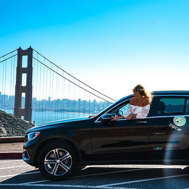 person leaning out of driver's side window of a car with San Francisco's Golden Gate Bridge in background