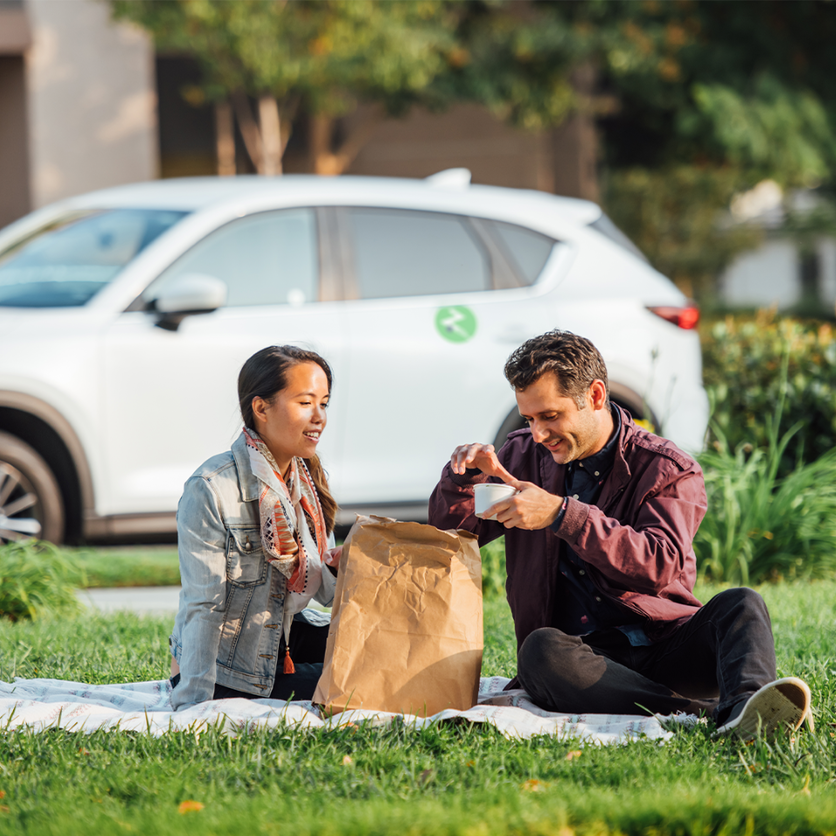 man and woman having a picnic on grass in front of white suv with zipcar logo