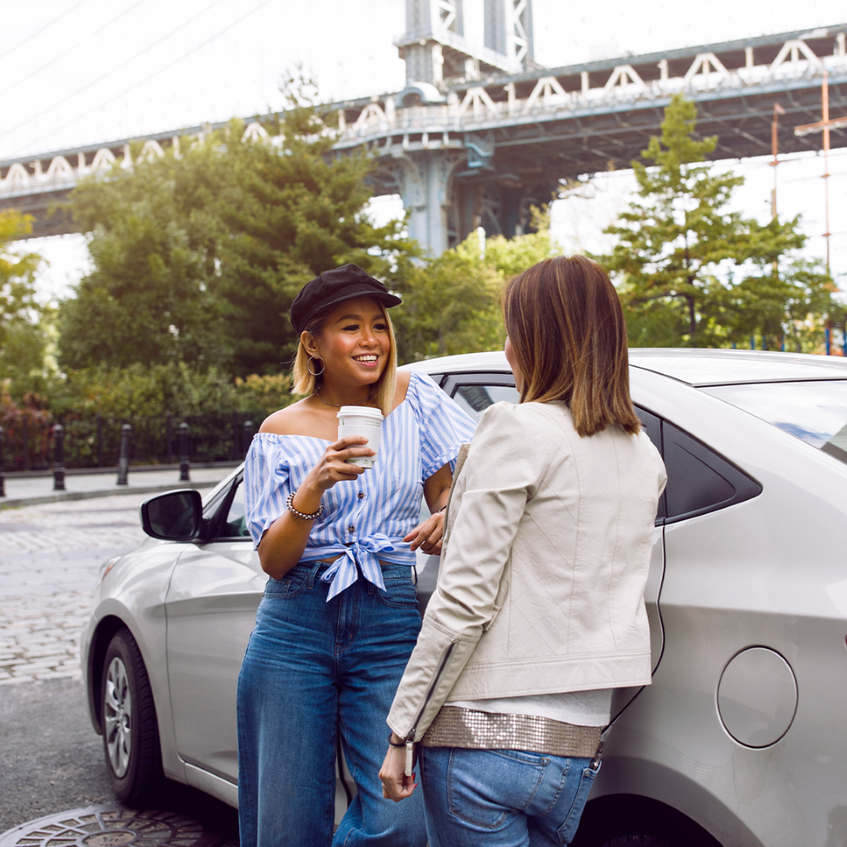 Two women leaning on a sedan and talking in the city near a bridge