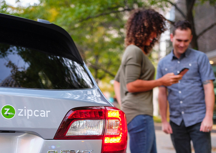 Woman showing a gentleman the Zipcar app while standing next to silver Zipcar.