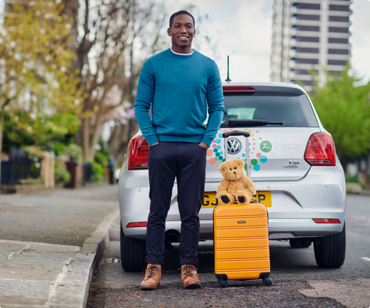 smiling man standing behind car with rolling suitcase